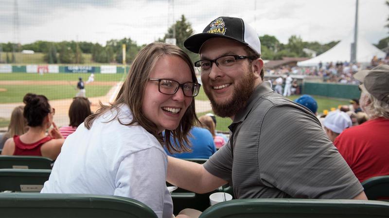 A man and woman smile at the camera with the baseball field in the background.